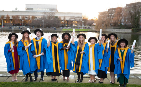 UCD graduates in blue doctoral robes stand together by the campus lake, smiling and holding their degree.